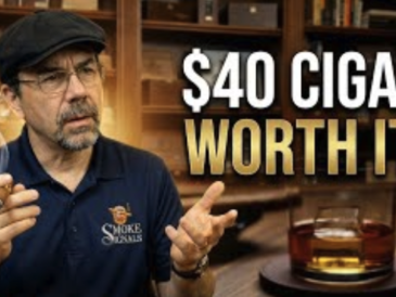 A man in glasses and a flat cap holds a smoking cigar, looking thoughtful. He wears a Smoke Signals shirt. Next to him, bold text reads $40 Cigar: WORTH IT? In front, a glass of whiskey sits on a table; shelves and cigars are in the background.